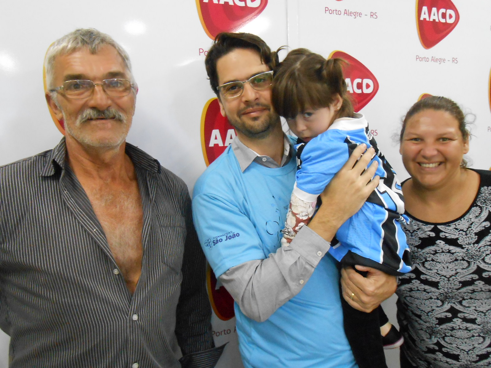 Carla tem três anos e faz tratamento na AACD. Estava na fila desta instituiçao para receber sua camiseta e finalmente chegou a sua vez para a alegria da sua mãe e avó que presenciaram a ação. E coube ao voluntário Ricardo Magrisso fazer a entrega do presentão azul.